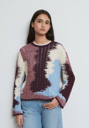 Joven con cabello largo y oscuro que lleva un suéter tejido multicolor en tonos burdeos, rosa, crema y azul, combinado con vaqueros azul claro.
