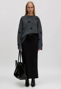 Grauer Strickpullover mit schwarzen Schleifenakzenten, kombiniert mit einem schwarzen langen Rock, einer glänzenden schwarzen Tote-Bag und spitzen schwarzen Stiefeletten.