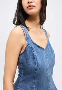 Νεαρή γυναίκα που φορεί αμάνικο μπλε τζιν φόρεμα, εμφανιζόμενη από τους ώμους μέχρι τη μέση, πάνω σε απλό φωτεινό φόντο.