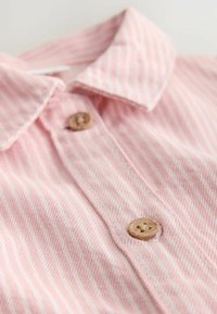 Jasnoróżowa koszula z kołnierzykiem w białe paski. Zawiera drewniane guziki i teksturowaną tkaninę. Zbliżenie podkreśla detale kołnierza i guzików.