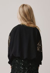 Črna jopa s prosto prilegajočimi se rokavi, okrašena s kristalčki na rokavih, z okroglim izrezom, kombinirana s črno krilo z reliefnimi kristalčki.