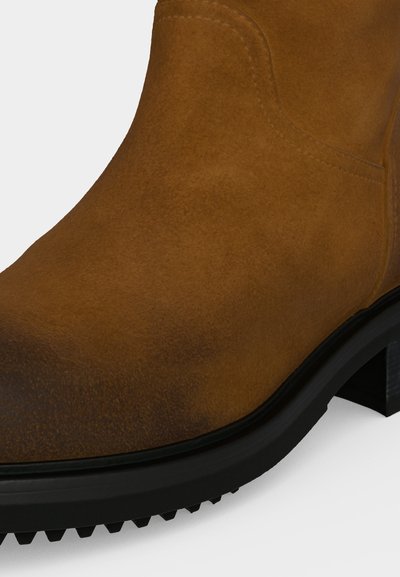 Gros plan d'une botte en daim marron avec semelle en caoutchouc noir et talon bas sur un fond clair.
