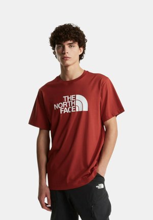 Giovane uomo che indossa una maglietta rossa The North Face e pantaloni neri con un logo bianco, in piedi con una mano in tasca.