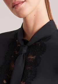Blusa nera con bordo in pizzo e dettaglio collo annodato; texture liscia con motivo floreale in pizzo intricato sulla parte anteriore.