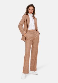 Brauner und orangefarbener karierter Anzug mit weiten Hosen und einem taillierten Blazer, kombiniert mit einer weißen Bluse und weißen Turnschuhen. Glatte Stofftextur.