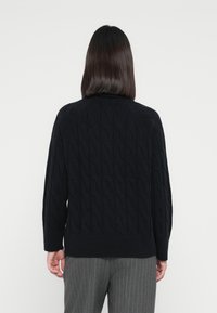 Maglione nero a maglia con motivo a treccia, polsini e orlo a coste. Indossato su pantaloni a righe grigi, mostrando una vestibilità rilassata.