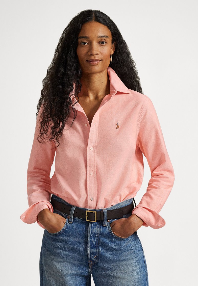 Gaiši rozā garām piedurknēm krekls ar apkakli, uz krūts izšūts logotips, kopā ar zilām džinsa biksēm un melnu jostu.