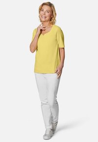 Geel T-shirt met korte mouwen en een V-halslijn, subtiele versieringen, gecombineerd met witte broek en lichtgrijze sneakers. Glad stoftextuur.