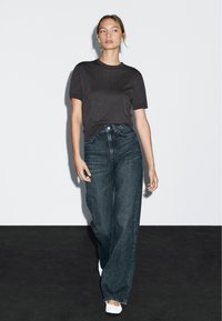 Tmavě šedé přiléhavé tričko v kombinaci s vysokými širokými džíny z tmavého denimu. Model má na sobě bílé slip-on boty. Jednoduché pozadí.