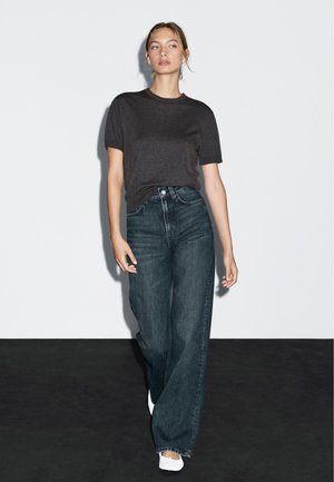 Tmavě šedé přiléhavé tričko v kombinaci s vysokými širokými džíny z tmavého denimu. Model má na sobě bílé slip-on boty. Jednoduché pozadí.