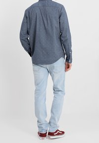 Camisa de manga larga azul marino con pequeños puntos blancos, combinada con jeans azul claro y zapatillas rojas. La vista trasera muestra el diseño ajustado de la camisa.