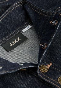 Tamsiai mėlynas džinsinis marškinis su kontrastinga oranžine siūle, juoda etikete su užrašu "JUXX" ir metaliniais snap užtrauktukais. Vidinė pamušalo dalis turi pilkas juostas.