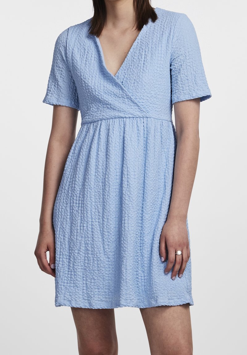 Femme portant une robe bleu clair texturée à manches courtes, col en V et taille froncée, debout devant un fond uni.