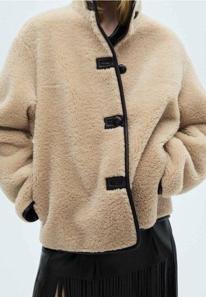 Femme portant une veste beige en peluche boutonnée avec une bordure noire et une jupe noire avec des détails à franges. Mains dans les poches de la veste.