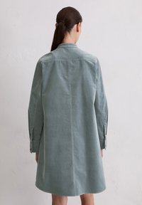 Vrouw met donker haar in een paardenstaart, gekleed in een losse, lange mouwen, gedempte groene jurk, die tegen een licht gestructureerde muur staat.