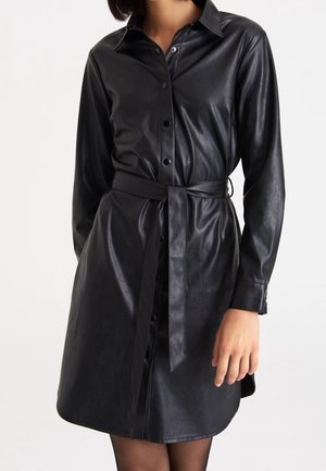 Schwarzes Kunstlederkleid mit Knöpfen, langen Ärmeln, Kragen und passendem Taillengürtel, getragen von einer Person vor einfarbigem Hintergrund.