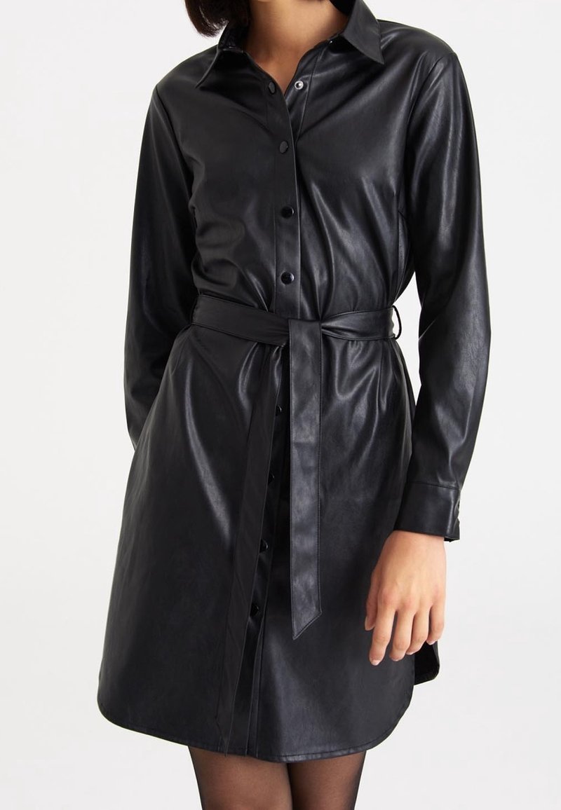 Robe noire en cuir synthétique boutonnée, manches longues, col et ceinture nouée assortie à la taille portée par une personne sur un fond uni.