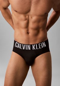 Torso maschile che indossa slip neri Calvin Klein, con addominali visibili e mani appoggiate sulla parte bassa della schiena su uno sfondo grigio neutro.