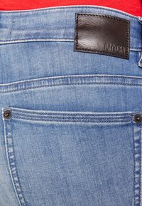 Jeans de mezclilla azul claro con un parche de cuero negro que lleva el texto "DRYKORN", costuras visibles y un acento de botón de metal en el bolsillo trasero.
