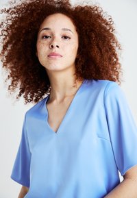 Lyseblå, kortermet bluse med V-halsdesign. Jevn tekstur med en subtil glans og en liten lomma på venstre side.