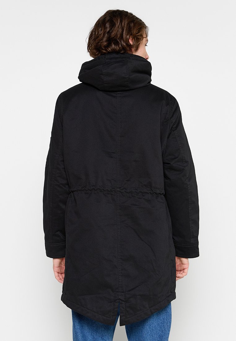 Veste noire à capuche avec taille resserrée, manches longues et dos légèrement allongé. Tissu texturé avec des détails de couture visibles.