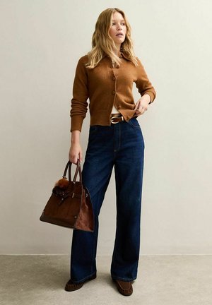 Cardigan de punto marrón, blusa blanca, jeans de pierna ancha de cintura alta en azul oscuro y bolso de ante marrón con un pompón. Zapatos planos marrones.