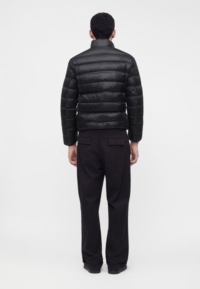 Homme debout, de dos, portant une veste matelassée noire, un pantalon noir avec des poches arrière et des chaussures noires sur un fond clair uni.