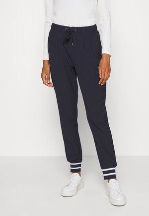 Pantalon de jogging noir avec taille à cordon et poignets rayés, porté avec des baskets blanches et un haut blanc à manches longues, montré de la taille vers le bas.