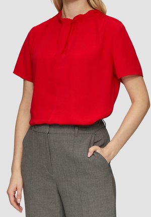 Femme portant un chemisier rouge vif à manches courtes rentré dans un pantalon gris taille haute et ajusté, avec une main dans la poche.