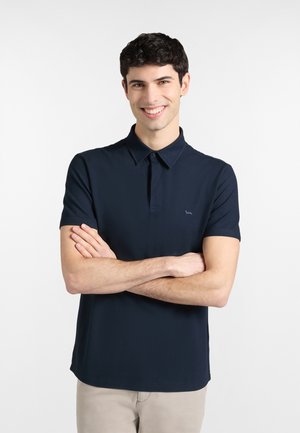 Giovane uomo sorridente con capelli corti e scuri, indossa una polo blu navy e pantaloni beige, con le braccia incrociate, in piedi davanti a uno sfondo bianco.