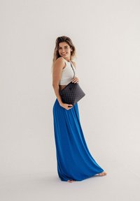 Černá látková kabelka s geometrickým vzorem, nošená přes tělo. Modelka v bílém tílku a modré maxi sukni, naboso, usmívající se.