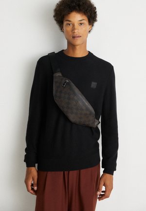 Jeune personne portant un pull noir et un pantalon marron avec un sac à bandoulière à carreaux foncés sur un fond clair uni.