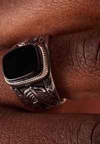 Zilveren ring met een rechthoekige zwarte steen, versierd met complexe bloemmotieven en gravures op de band, gepresenteerd op een gestructureerde huid.