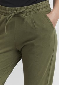Person som har på seg olivengrønne sweatpants med snor i livet og hånd i høyre lomme.