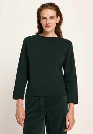 Woman wearing dark green long-sleeve sweater with folded cuffs and matching corduroy pants, standing against a plain light background.
