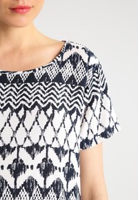 Kortärmad topp med ett marinblått och vitt abstrakt mönster, bred halsringning och mjuk textur, som betonar geometriska former och djärva mönster.