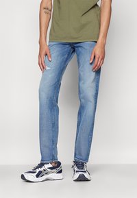 Jeans rectos de color azul claro con un sutil desgaste y detalles rasgados en la rodilla, combinados con una camiseta holgada de color verde oliva y zapatillas blancas.