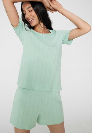 Mujer sonriente con cabello largo y oscuro, lleva una camiseta de manga corta de punto verde claro y pantalones cortos de punto a juego, posando con los brazos levantados frente a un fondo liso.