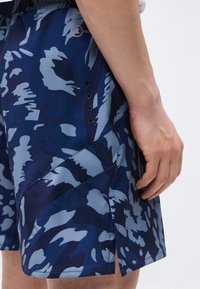 Ανδρικό αθλητικό σορτς σε μπλε και γκρι καμουφλάζ, με τρύπες αερισμού στο πλάι και σκίσιμο στο τελείωμα, φορεμένο από άτομο που στέκεται με το χέρι χαλαρό.