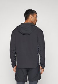 Chaqueta negra con capucha de diseño ajustado, textura suave y mangas largas, llevada sobre pantalones cortos a juego, vista desde la parte de atrás.