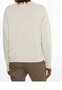 Beżowy sweter dzianinowy z prążkowanym dekoltem i mankietami, z wąskim paskiem wzdłuż pleców. Noszony z brązowymi spodniami w kratę.