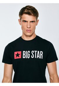 Młody mężczyzna z krótkimi brązowymi włosami, ubrany w czarną koszulkę z czerwonym gwiazdką i napisem „BIG STAR” na jednolitym białym tle.