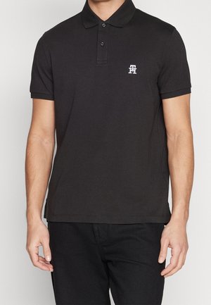 Hombre con camiseta tipo polo negra de manga corta con un pequeño logo blanco bordado en el pecho izquierdo, combinado con pantalones negros.