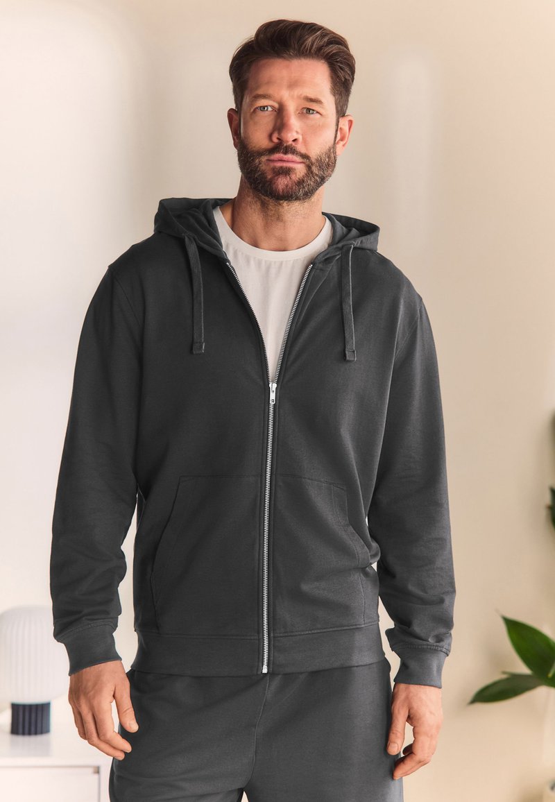 Mężczyzna z brodą w czarnej bluzie na suwak i białym t-shircie, stoi w pomieszczeniu na neutralnym tle, w pobliżu meble i roślina.