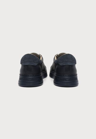 Paire de baskets en cuir noir avec languettes de talon en tissu foncé, vues de dos sur un fond clair uni.