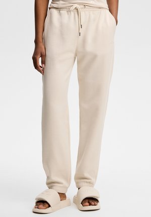Person trägt beige Sweatpants mit Seitentaschen und passenden beigen Slide-Sandalen auf weißem Hintergrund.