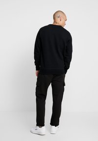 Schwarzer Pullover mit Rundhalsausschnitt, kombiniert mit schwarzen Cargo-Hosen mit seitlichen Taschen und weißen Sneakers auf einem hellen Hintergrund.