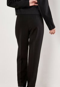 Een persoon in een zwart op maat gemaakte broek en een bijpassende zwarte blazer, met één hand die de blazer aan de achterkant aanpast.