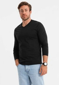 Maglietta nera a maniche lunghe con scollo a V, realizzata in tessuto morbido, abbinata a jeans azzurri chiari. Il modello indossa un orologio nero al polso.