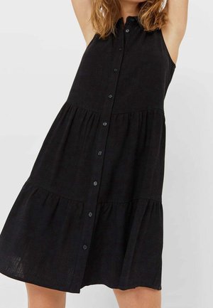 Sleeveless black button-down dress with tiered skirt worn by person with raised arms against plain background.